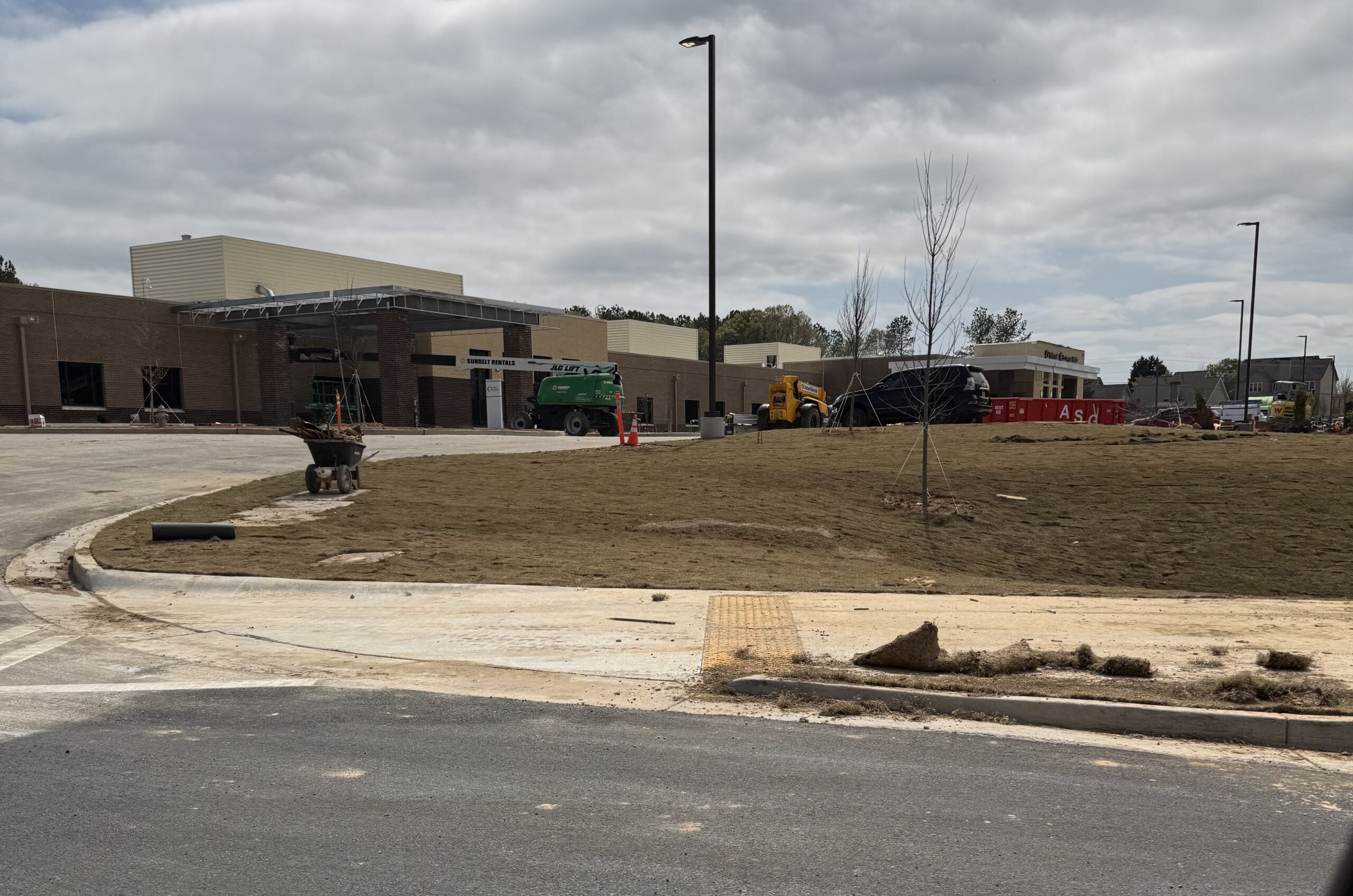 Front entrance and main driveway of Piedmont Encompass Health Eastside Rehabilitation Hospital under construction — curbed drive approach, young trees planted, equipment and workers visible, Loganville GA 2026